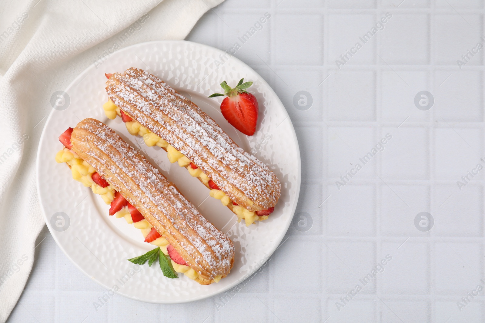 Photo of Delicious eclairs filled with cream, strawberries and mint on white tiled table, top view. Space for text