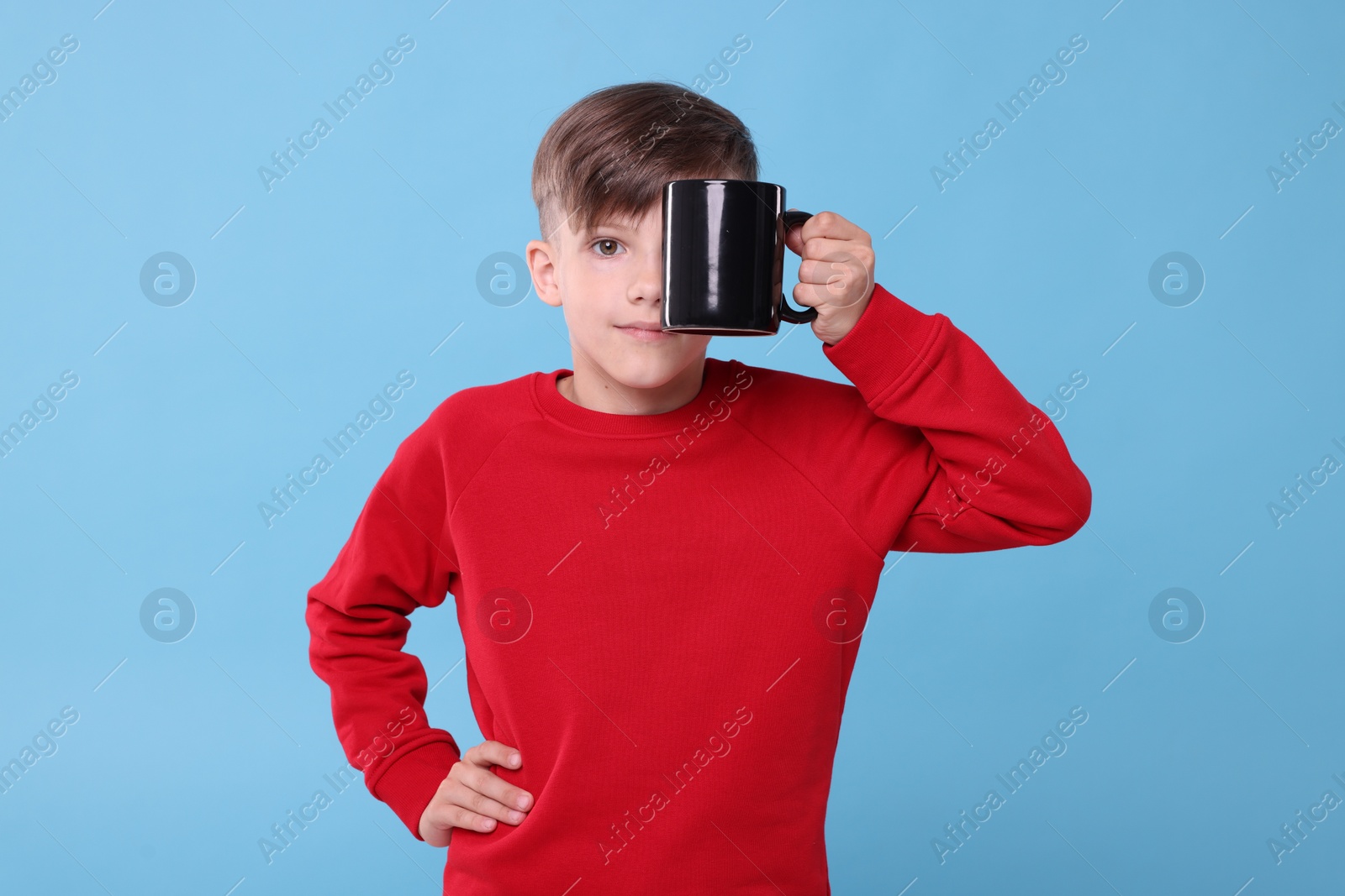 Photo of Cute boy covering eye with black ceramic mug on light blue background