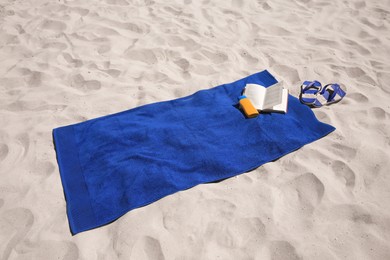 Photo of Soft blue beach towel, flip flops, sunblock and book on sand