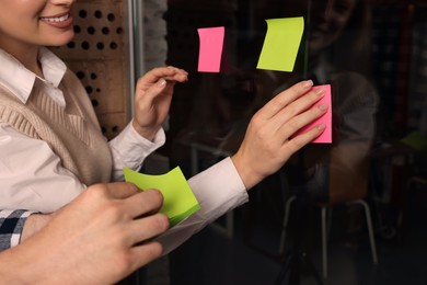 Photo of Employees putting colorful sticky notes on glass door in office, closeup. Team work motivation