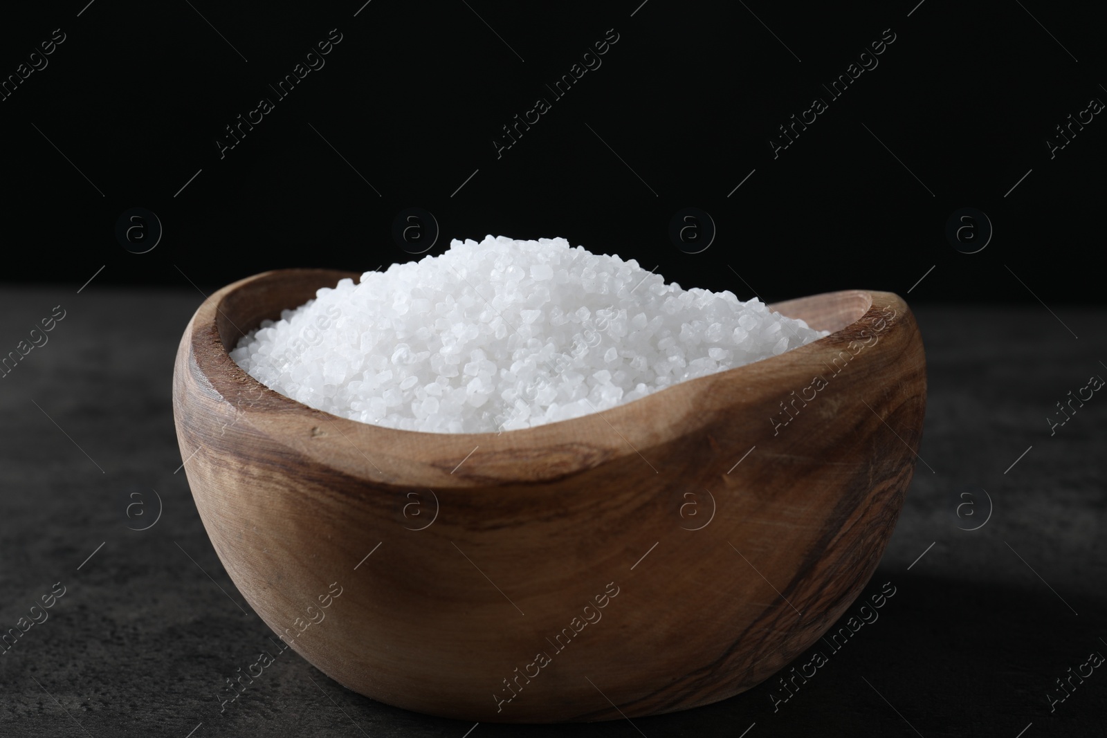 Photo of Natural salt in wooden bowl on dark grey table, closeup