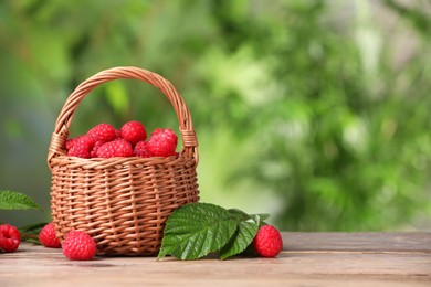 Wicker basket with tasty ripe raspberries and leaves on wooden table against blurred green background, space for text