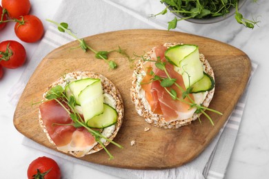 Photo of Crunchy buckwheat cakes with cream cheese, prosciutto and cucumber slices on white marble table, flat lay