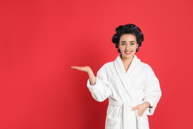 Photo of Happy young woman in bathrobe with hair curlers on red background, space for text