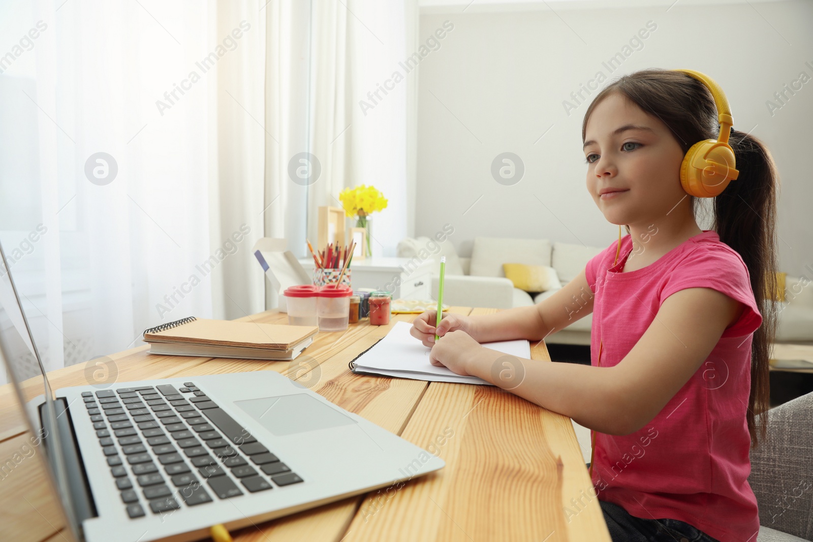 Photo of Little girl with headphones drawing on paper at online lesson indoors. Distance learning