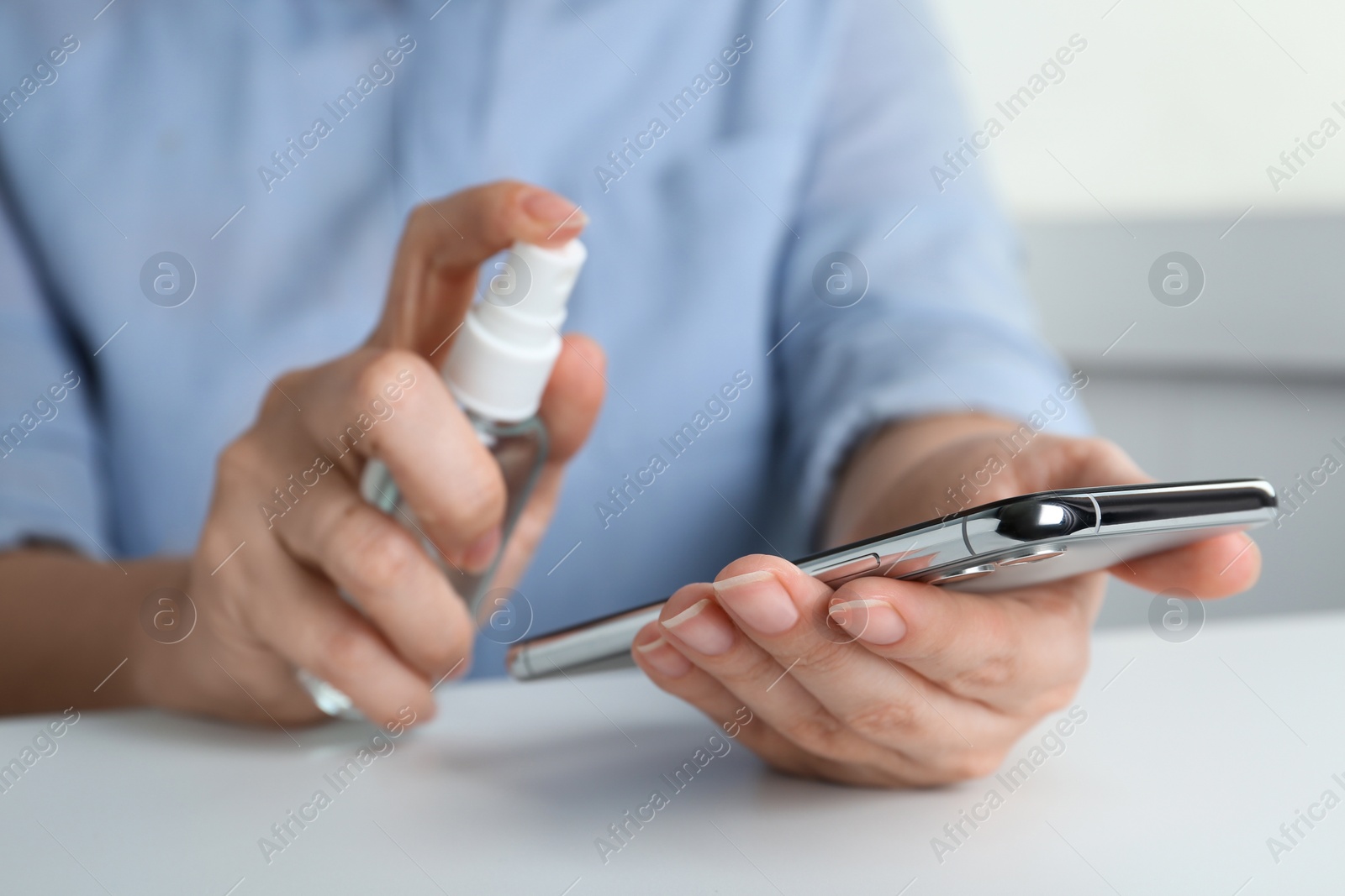 Photo of Woman spraying antiseptic onto smartphone at white table, closeup