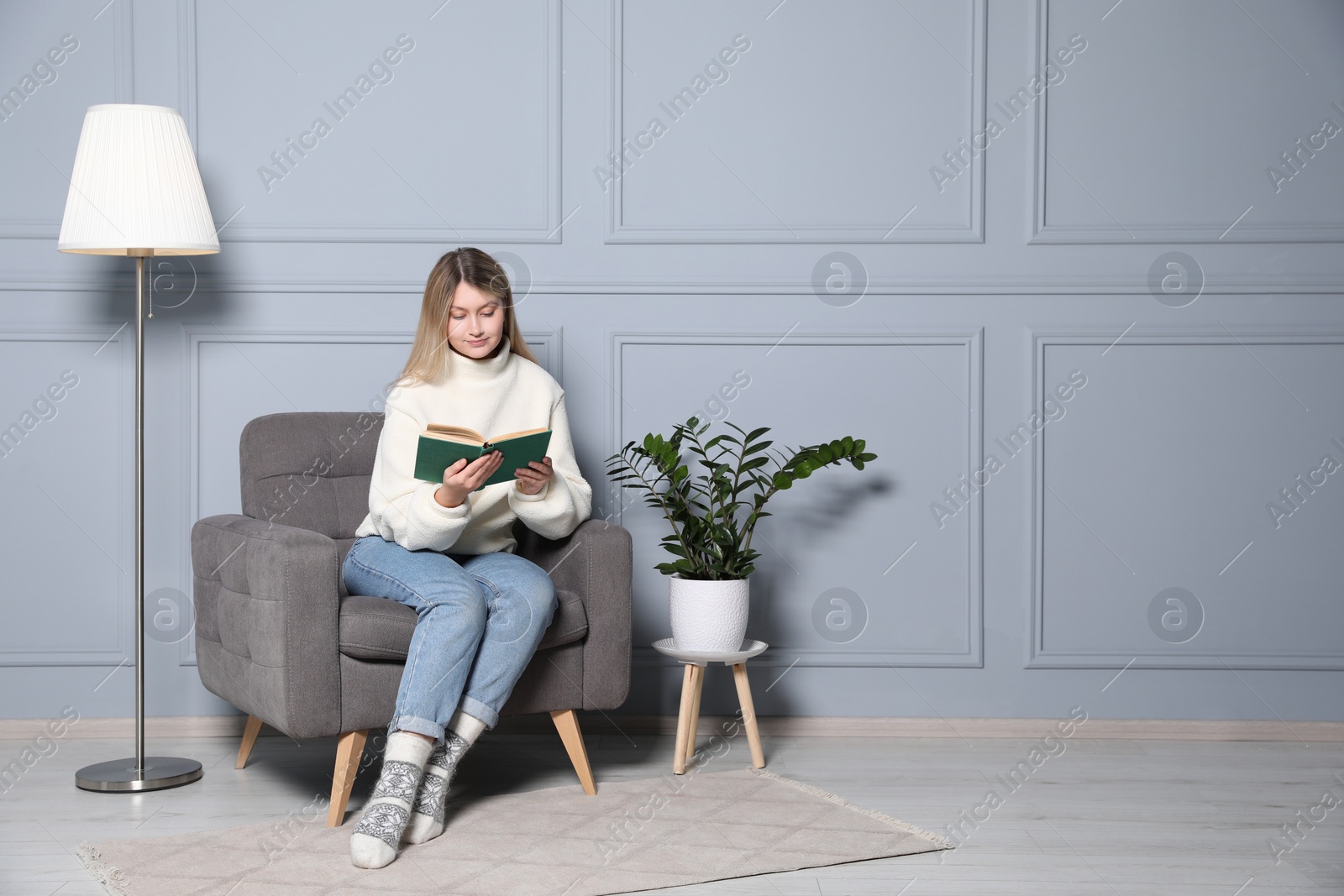 Photo of Young woman reading book in armchair at home, space for text. Interior design