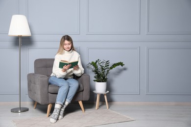 Young woman reading book in armchair at home, space for text. Interior design