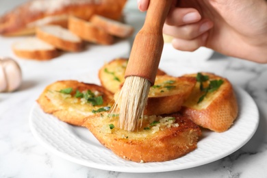 Photo of Woman brushing slices of delicious toasted bread with garlic and herbs on white marble table, closeup