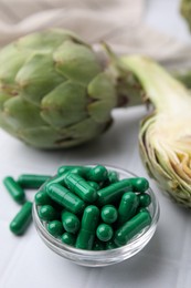 Photo of Bowl with pills and fresh artichokes on white tiled table, closeup