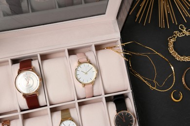 Jewelry box with stylish wristwatches and golden accessories on black table, flat lay
