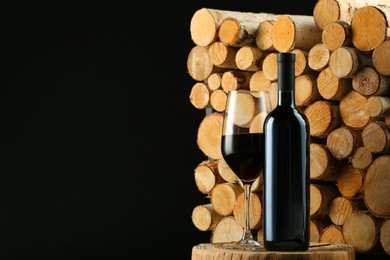 Stylish presentation of red wine in bottle and wineglass near wooden logs on black background, space for text