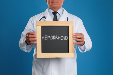 Doctor holding blackboard with word HEMORRHOID on blue background, closeup