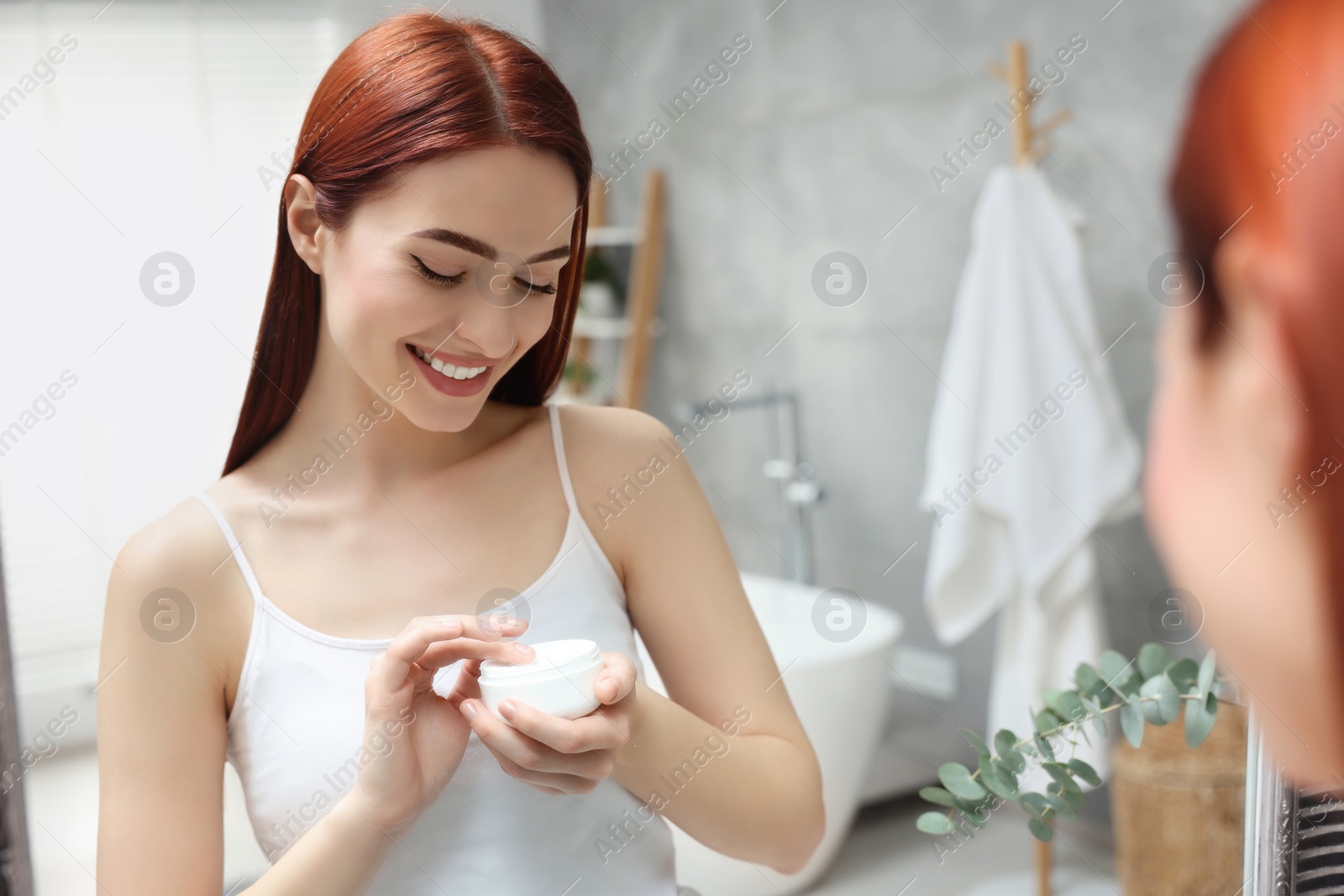 Photo of Beautiful young woman holding jar of body cream near mirror in bathroom