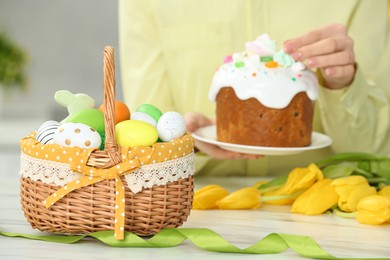 Photo of Closeup of Easter basket with painted eggs on white marble table near woman with cake, focus on food