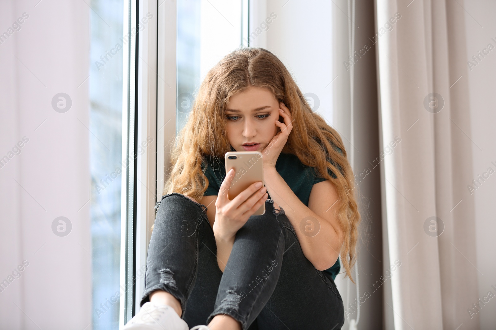 Photo of Upset woman with smartphone sitting on window sill indoors. Loneliness concept