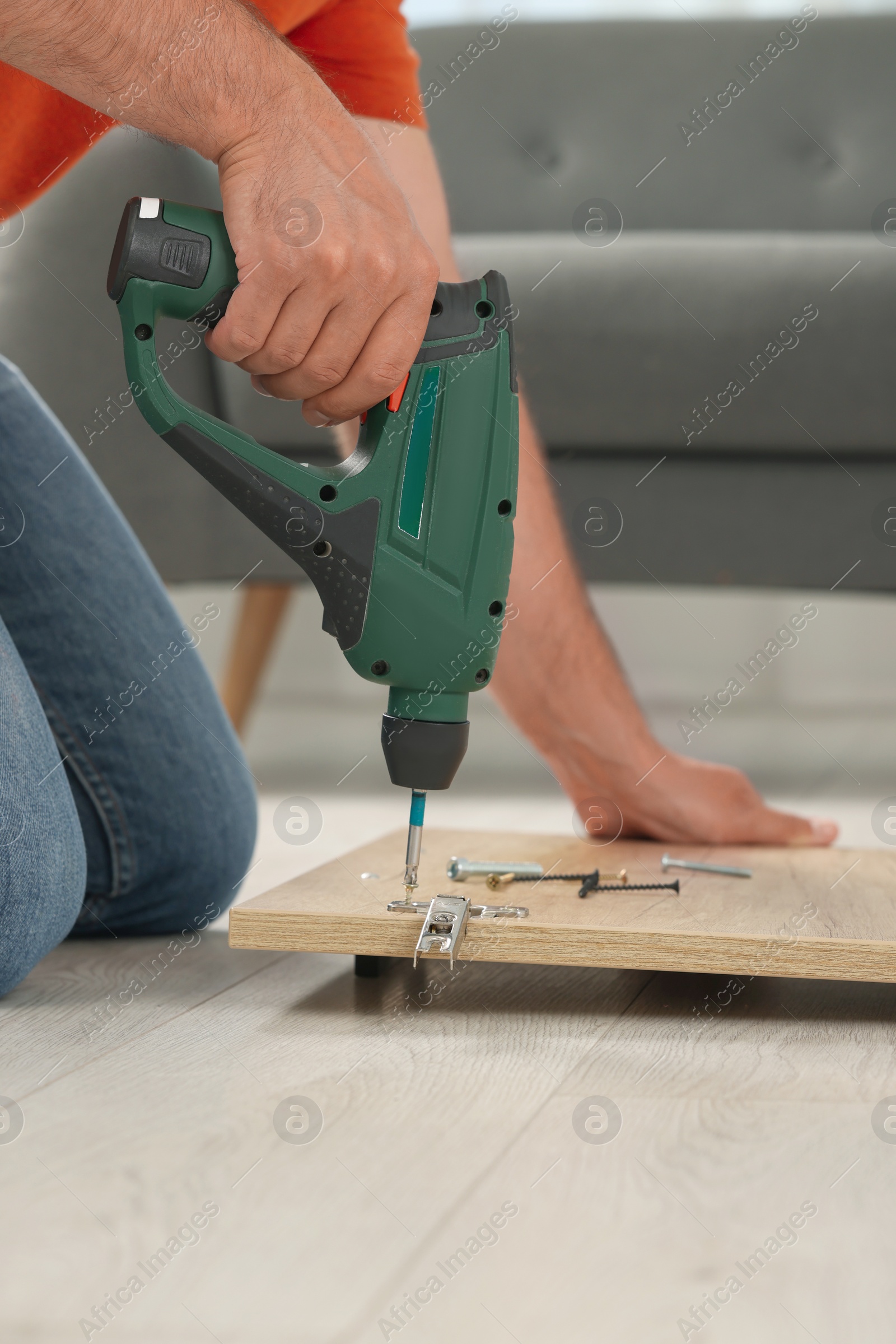 Photo of Man with electric screwdriver assembling furniture on floor indoors, closeup