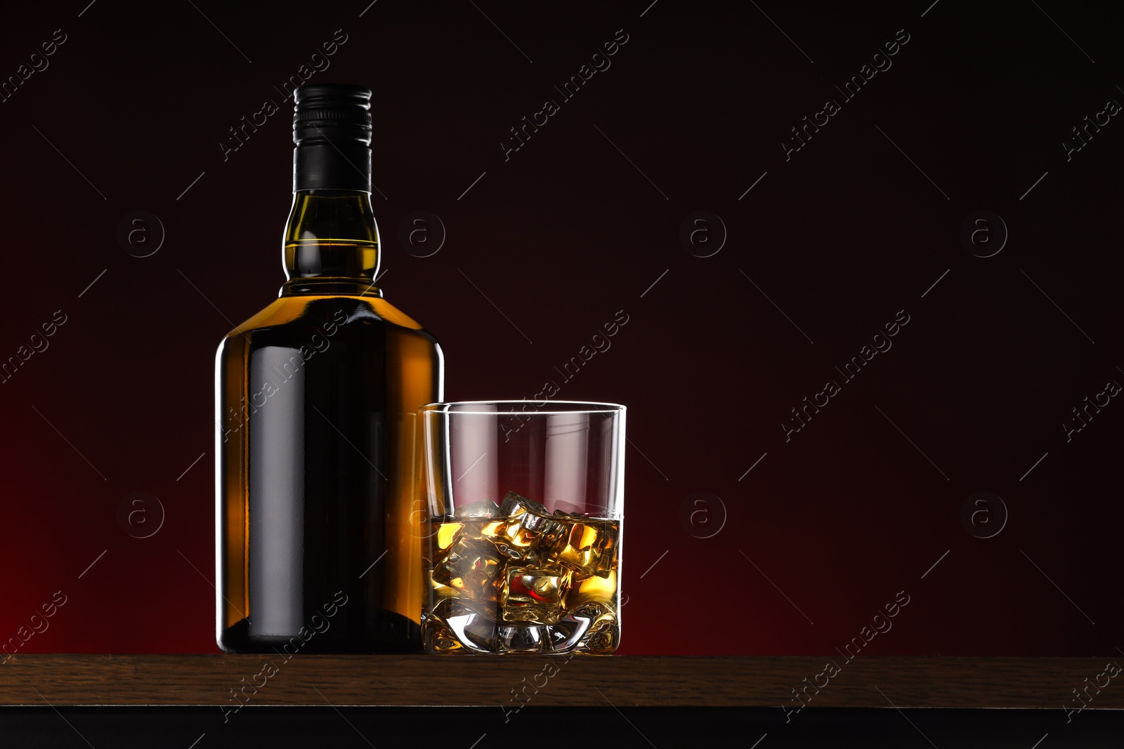 Photo of Whiskey with ice cubes in glass and bottle on wooden table, space for text