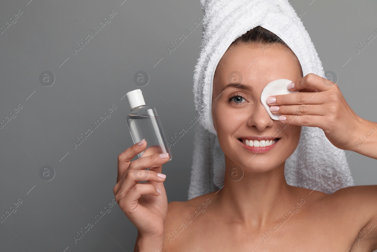 Photo of Smiling woman removing makeup with cotton pad and holding bottle on grey background. Space for text