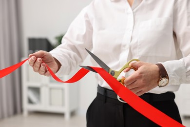 Photo of Woman cutting red ribbon with scissors indoors, closeup