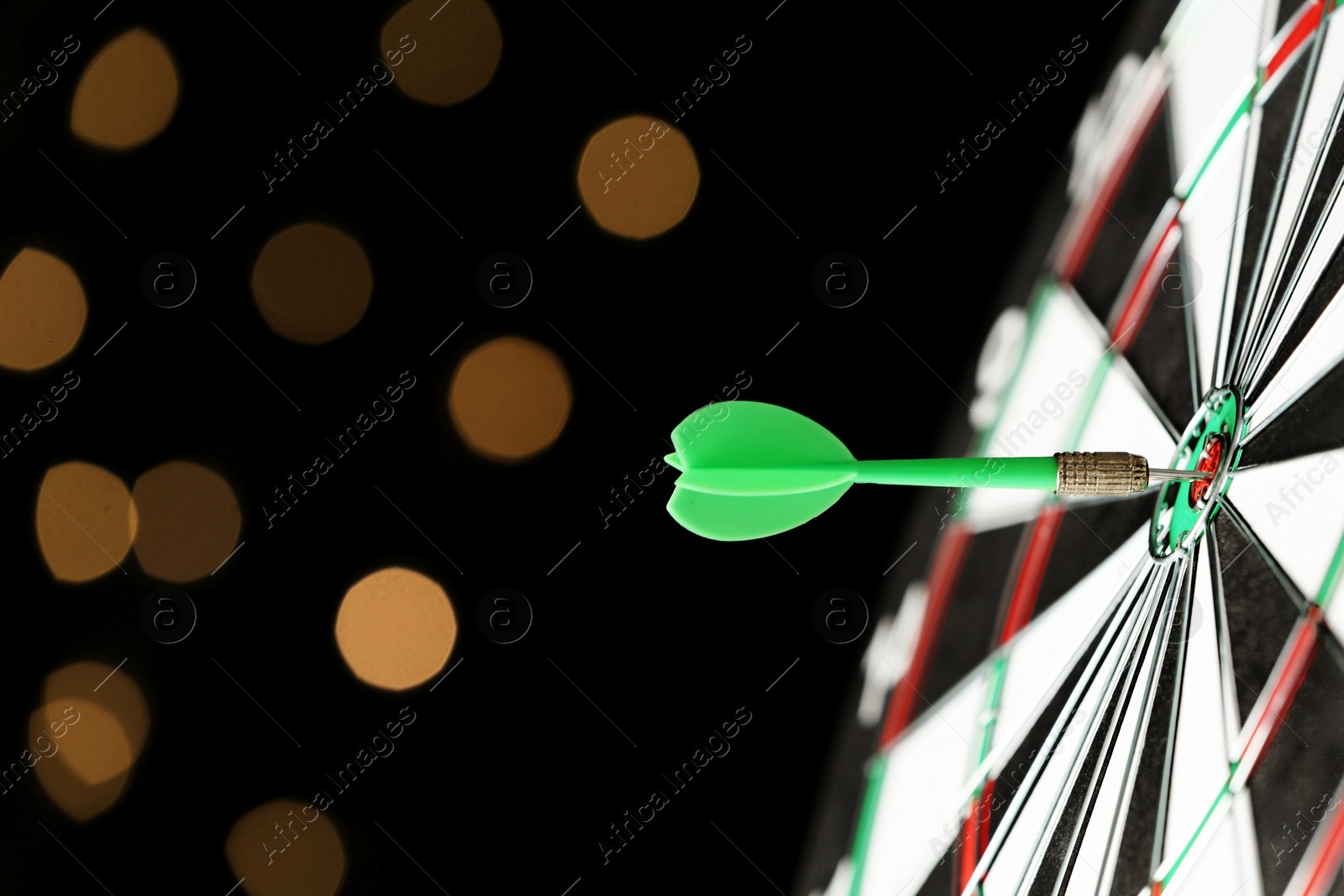 Photo of Green arrow hitting target on dart board against blurred lights. Space for text