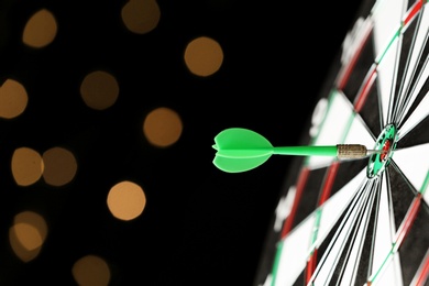 Photo of Green arrow hitting target on dart board against blurred lights. Space for text