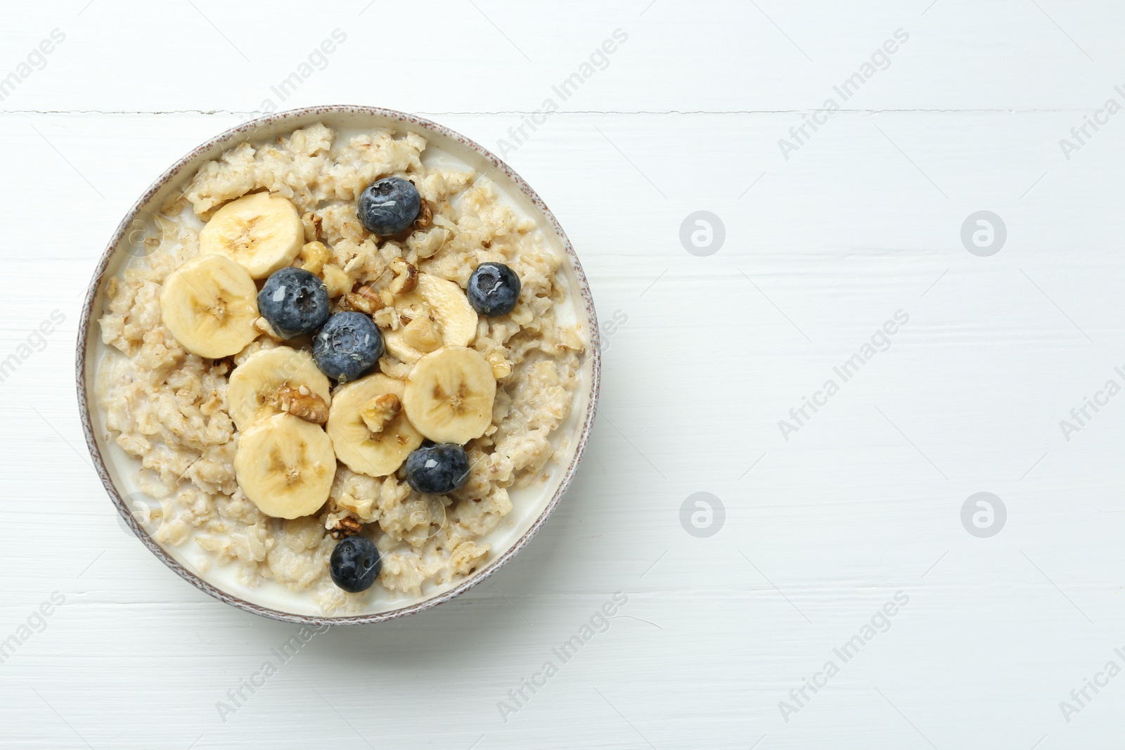 Photo of Tasty oatmeal with banana, blueberries, walnuts and honey served in bowl on white wooden table, top view. Space for text