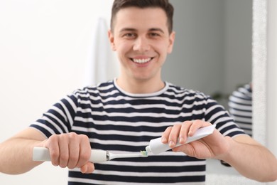 Photo of Man squeezing toothpaste from tube onto electric toothbrush in bathroom, selective focus