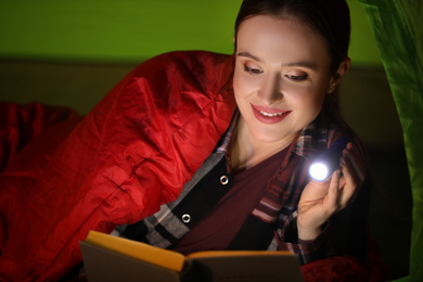 Young woman with flashlight reading book in tent