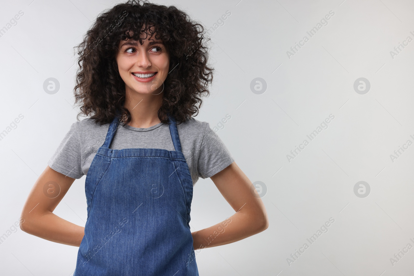 Photo of Happy woman wearing kitchen apron on light grey background, space for text. Mockup for design