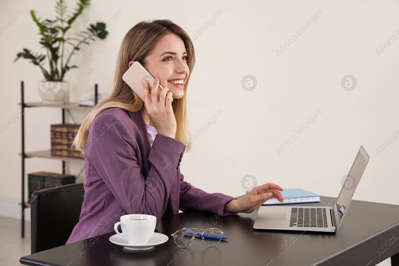 Image of Young woman talking on phone while using laptop in office