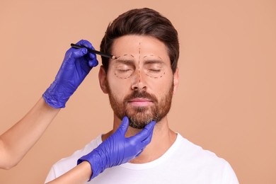 Doctor drawing marks on man's face for cosmetic surgery operation against beige background