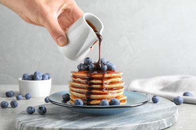 Photo of Woman pouring chocolate syrup onto fresh pancakes with blueberries at grey table, closeup