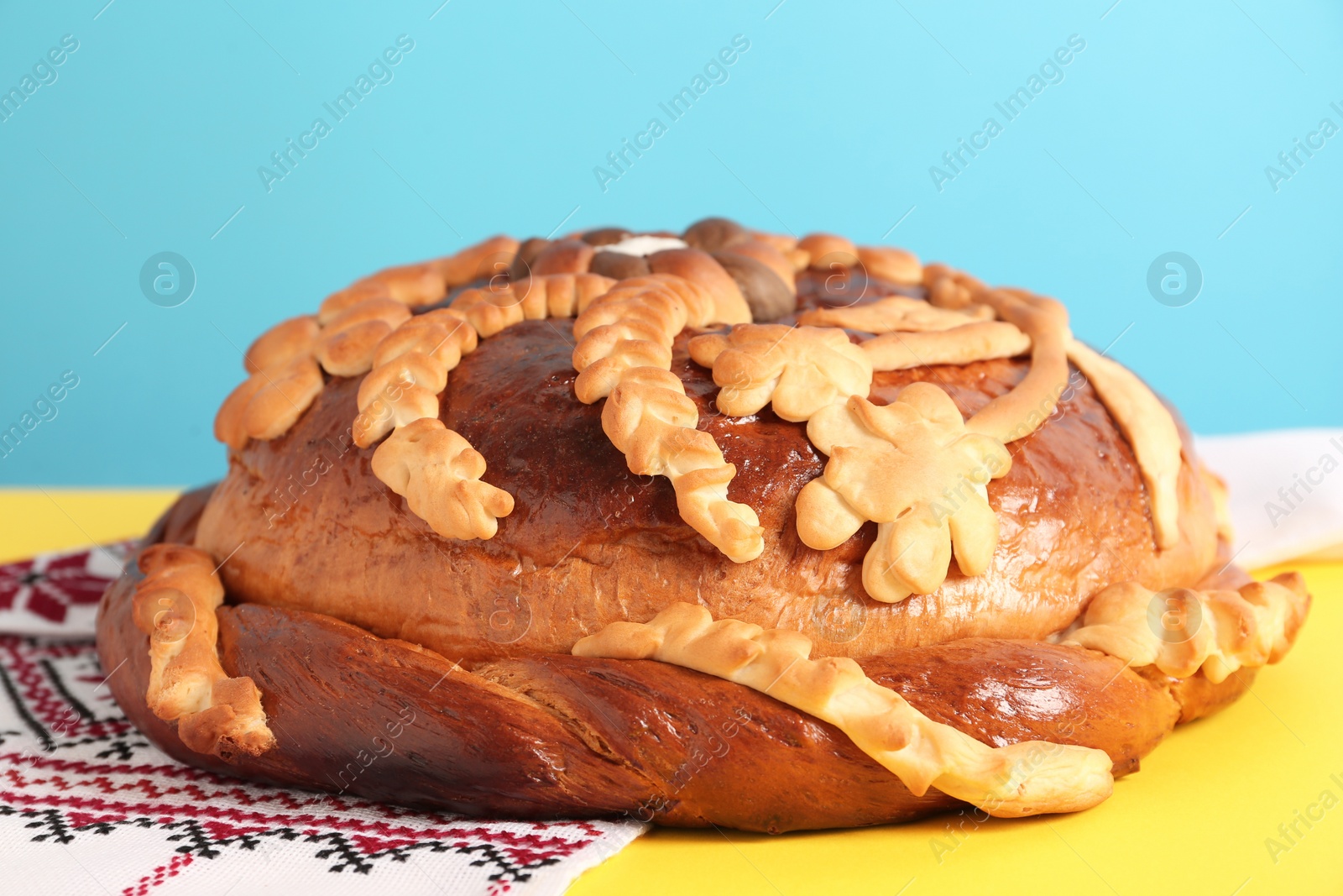 Photo of Rushnyk with korovai on yellow table against light blue background, closeup. Ukrainian bread and salt welcoming tradition