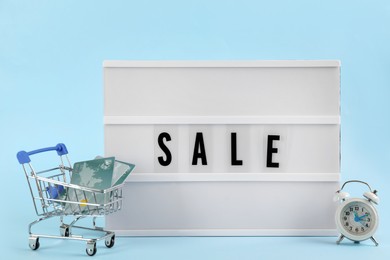 Photo of Lightbox with word Sale, alarm clock and credit cards in shopping cart on light blue background