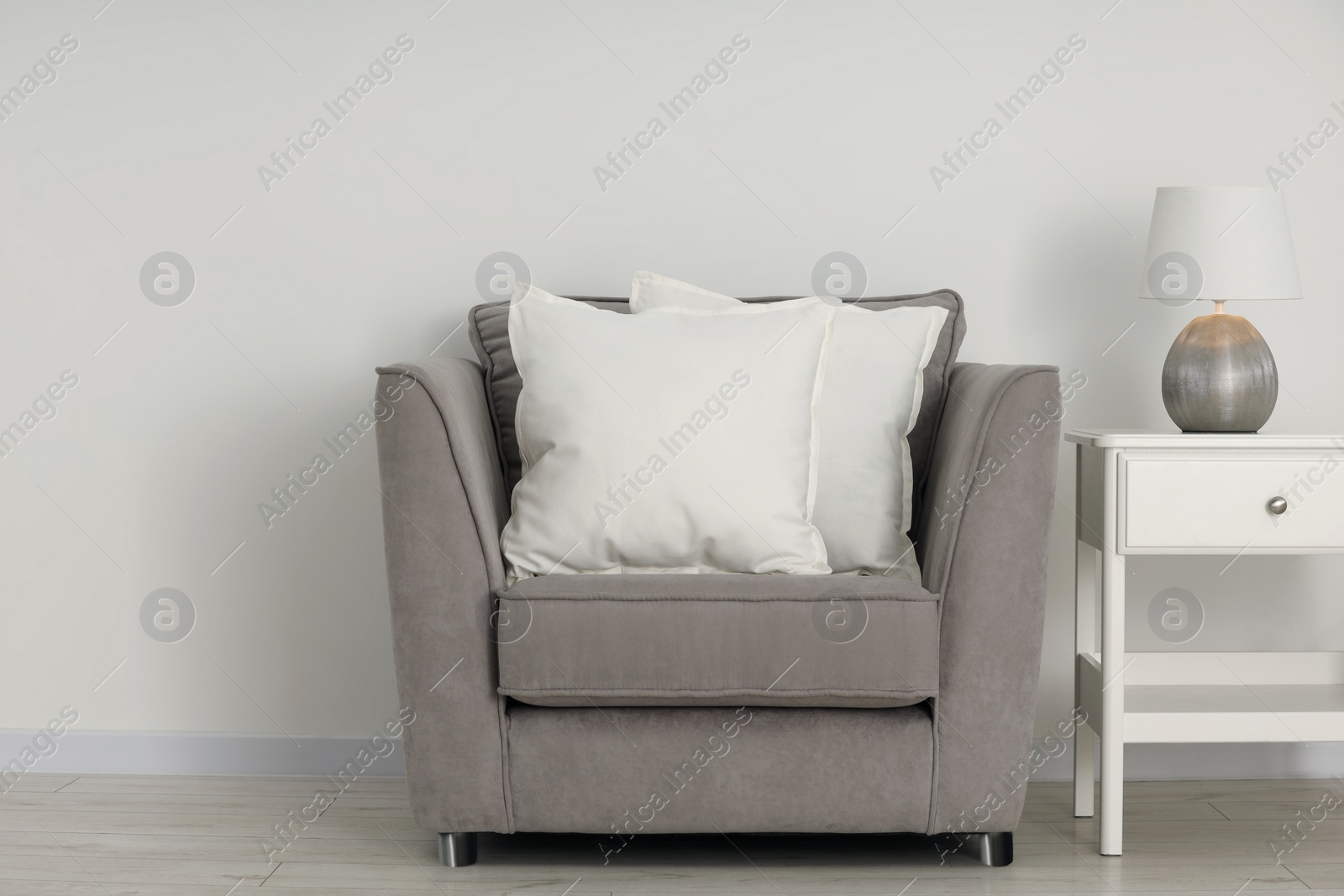 Photo of Soft white pillows on armchair, bedside table and lamp near light wall indoors