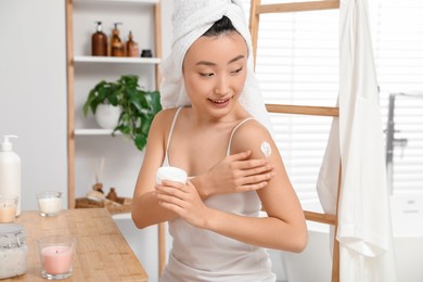 Photo of Beautiful young Asian woman applying body cream on shoulder in bathroom
