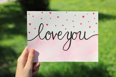 Photo of Woman holding card with phrase Love You outdoors, closeup