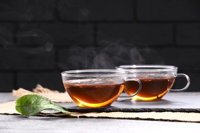 Photo of Aromatic hot tea in glass cups and green leaves on light grey table near brick wall