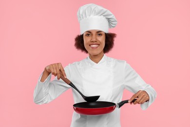 Photo of Happy female chef in uniform holding frying pan and ladle on pink background