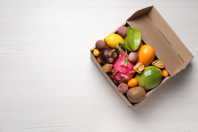 Photo of Cardboard box with different exotic fruits on white wooden table, top view. Space for text