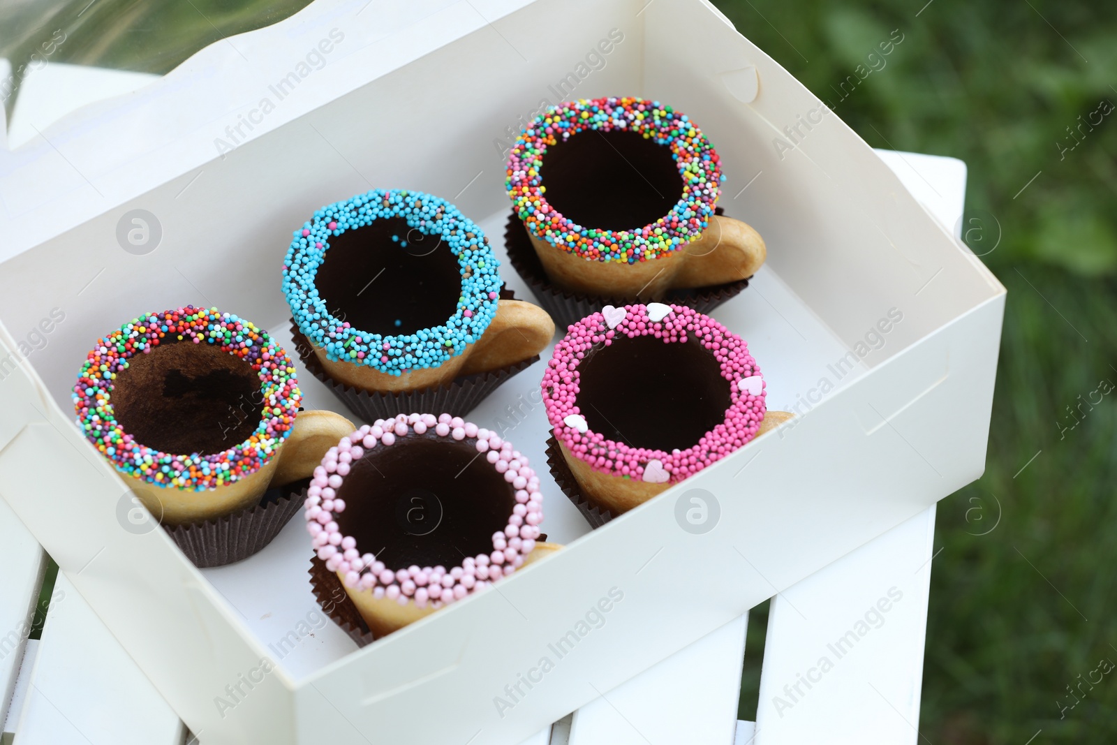 Photo of Box of delicious edible biscuit coffee cups decorated with sprinkles on white wooden table outdoors
