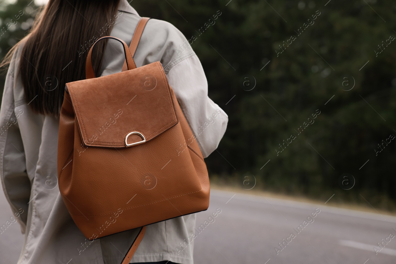 Photo of Young woman with stylish backpack near empty road in forest, back view. Space for text