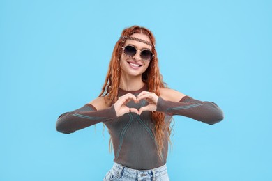 Photo of Stylish young hippie woman in sunglasses making heart with hands on light blue background