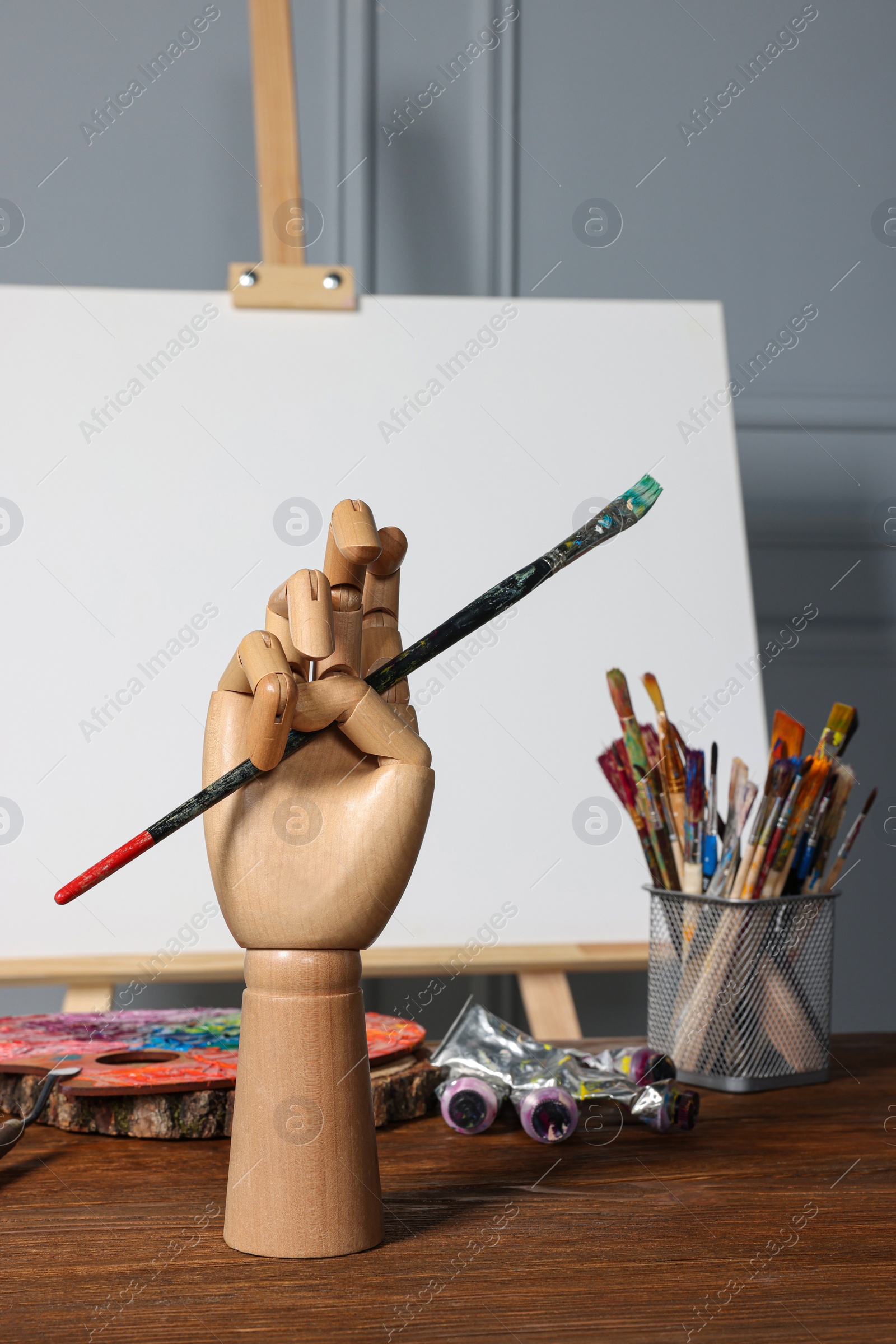 Photo of Easel with blank canvas, hand model and different art supplies on wooden table near grey wall