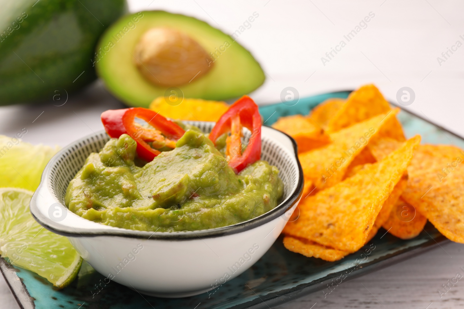 Photo of Bowl of delicious guacamole with chili pepper and nachos chips on white wooden table, closeup