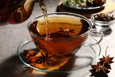 Pouring aromatic anise tea from teapot into glass cup on light grey table, closeup