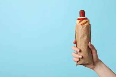 Woman holding paper bag with delicious french hot dog on light blue background, closeup. Space for text
