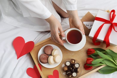 Photo of Tasty breakfast served in bed. Woman with tea, desserts, gift box and flowers at home, top view