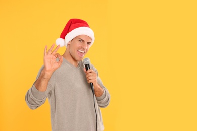 Photo of Emotional man in Santa Claus hat singing with microphone on yellow background, space for text. Christmas music